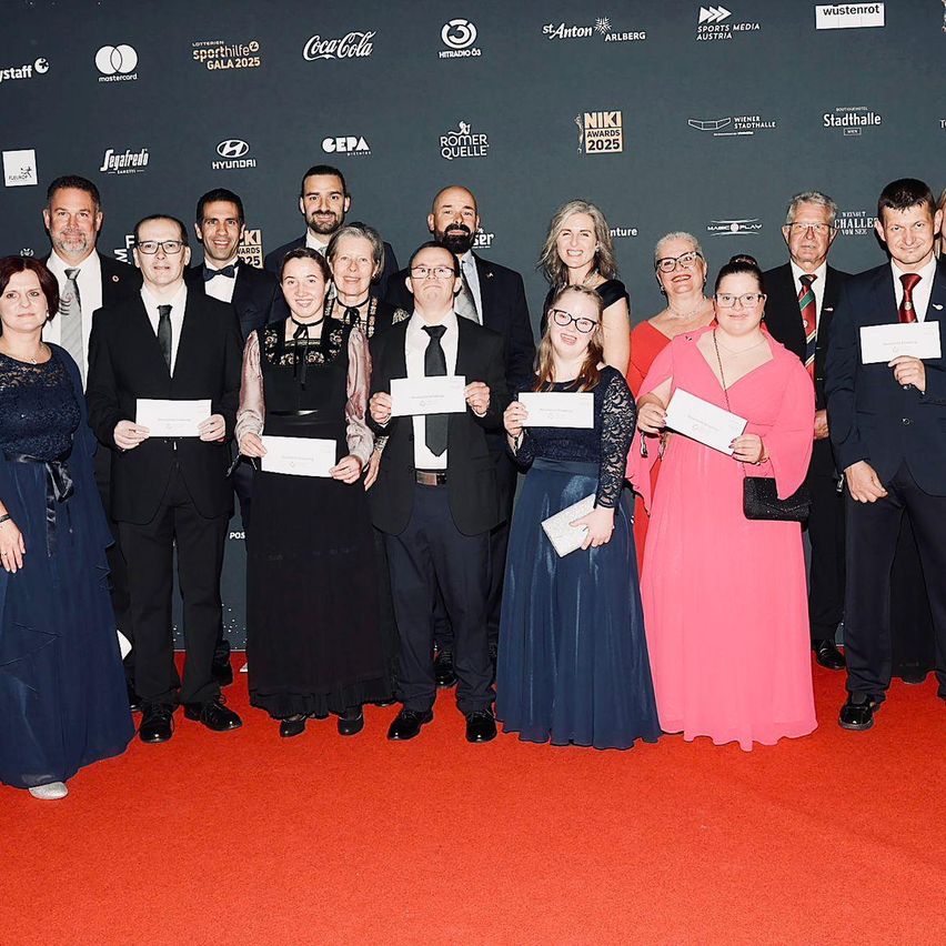 Eine Gruppe von Menschen in formeller Kleidung steht auf einem roten Teppich und hält Urkunden. Hinter ihnen befindet sich eine Wand mit Logos und Texten, darunter 'NIKI Awards 2025'. Sie lächeln für die Kamera.