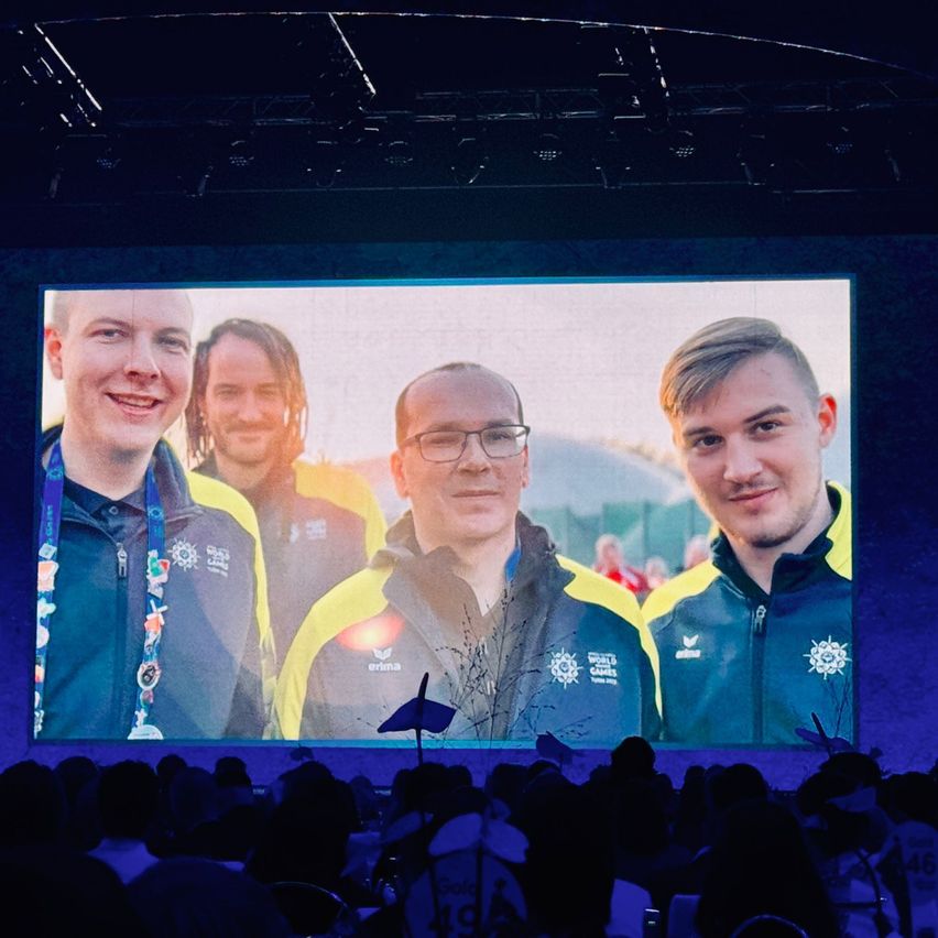 Eine Gruppe junger Männer mit Medaillen und Jacken lächelt in die Kamera. Sie stehen vor einer großen Leinwand, die ihr Bild zeigt. Das Publikum darunter beobachtet sie.