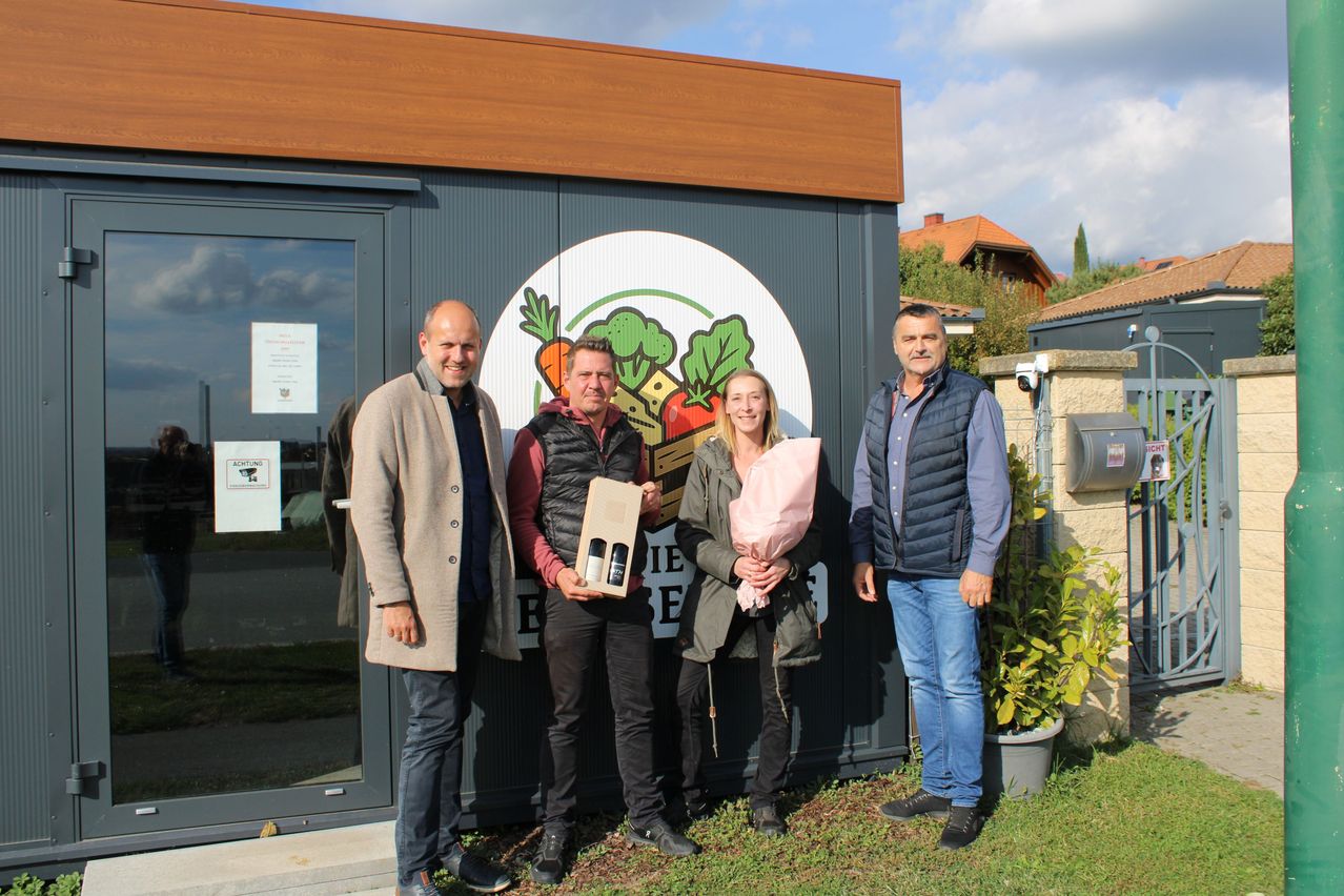 Vier Menschen stehen vor einem Gebäude, zwei Männer und zwei Frauen. Sie lächeln und scheinen für ein Foto zu posieren. Die Frau auf der rechten Seite hält einen Blumenstrauß. Das Gebäude hat eine Glastür und ein Schild mit einem Logo, das Gemüse zeigt.