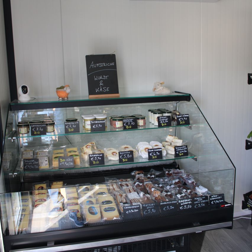 Eine Vitrine mit Glasregalen und einem Schild mit der Aufschrift Aufstriche Wurst & Käse zeigt verschiedene Lebensmittel wie Käse, Marmeladen und Gebäck. Einige Artikel haben Preisschilder. Die Umgebung scheint ein Geschäft oder ein Marktstand zu sein.