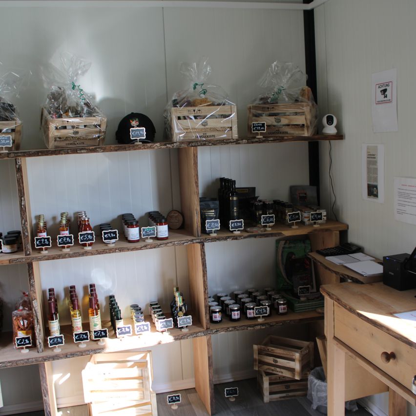 Ein hölzerne Regal mit verschiedenen Gläsern und Flaschen. Preisschilder sind an den Flaschen angebracht. Drei Holzkisten stehen auf dem obersten Regal. Ein Schreibtisch mit einem Mülleimer steht rechts. Eine Überwachungskamera ist an der Wand montiert.