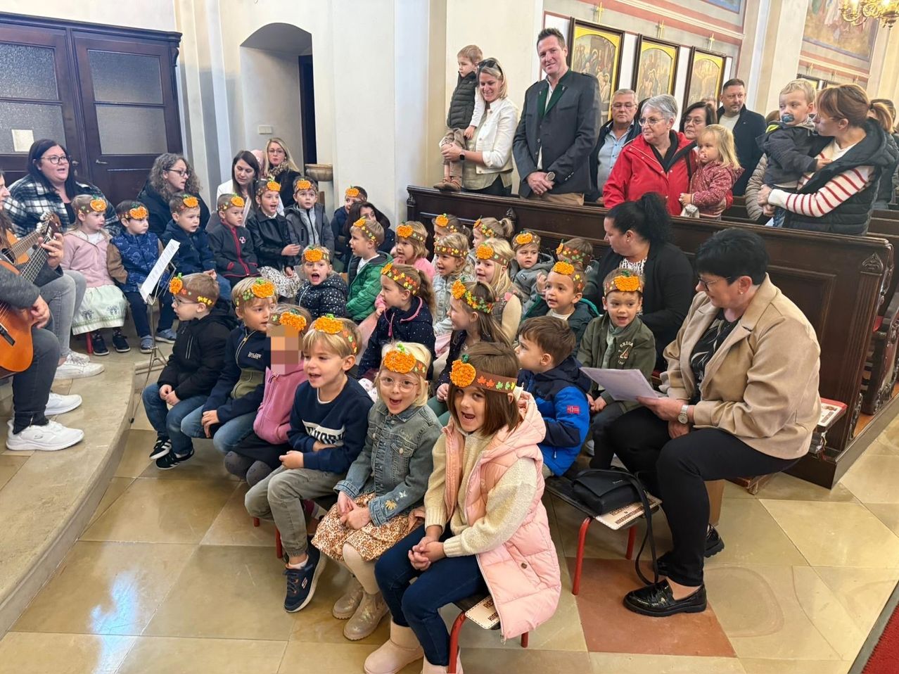 Eine Gruppe von Kindern mit Blumenkopfbändern sitzt in einer Kirche. Erwachsene stehen hinter ihnen, einige beobachten. Eine Frau sitzt und schreibt auf Papier.