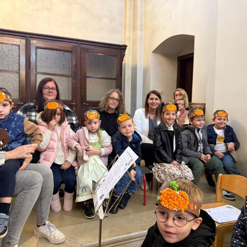 Eine Gruppe von Kindern mit Kopfbändern in Kürbisform sitzt in einem Raum zusammen, einige mit Musikinstrumenten. Erwachsene sitzen mit ihnen, und eine Frau hält ein Notenpult.