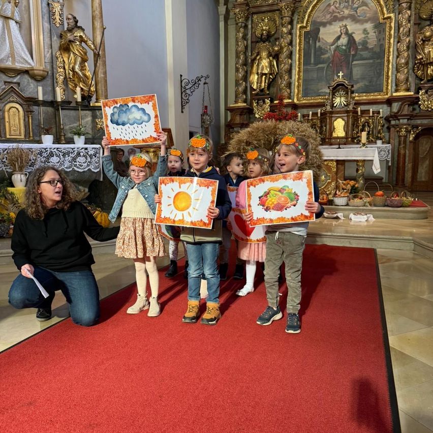 In einer Kirche kniet eine Frau und hält ein Papier, während Kinder auf einem roten Teppich stehen und Zeichnungen von Wetter und Früchten halten. Der Altar ist mit einem Gemälde geschmückt.