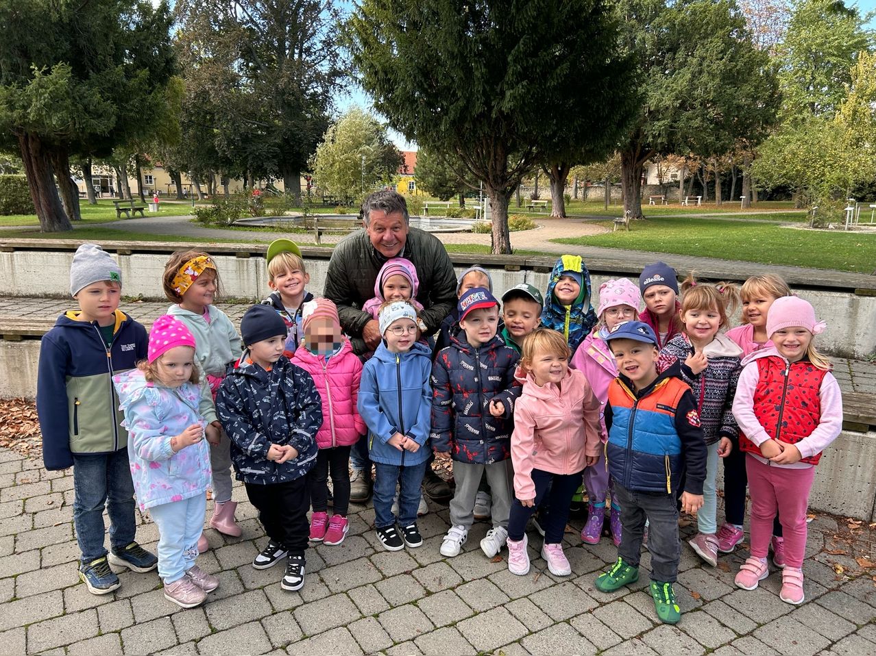 Eine Gruppe von Kindern und einem Mann stehen in einem Park und lächeln für ein Foto. Bäume und Bänke sind im Hintergrund zu sehen.