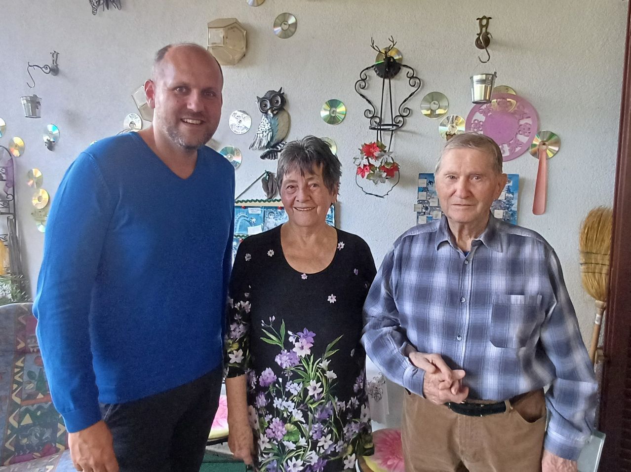 Drei Personen posieren für ein Foto in einem Raum mit Blumenarrangement, Eule und dekorativen Tellern. Ein Mann in Blau, eine Frau in Schwarz und ein älterer Mann in Karo.