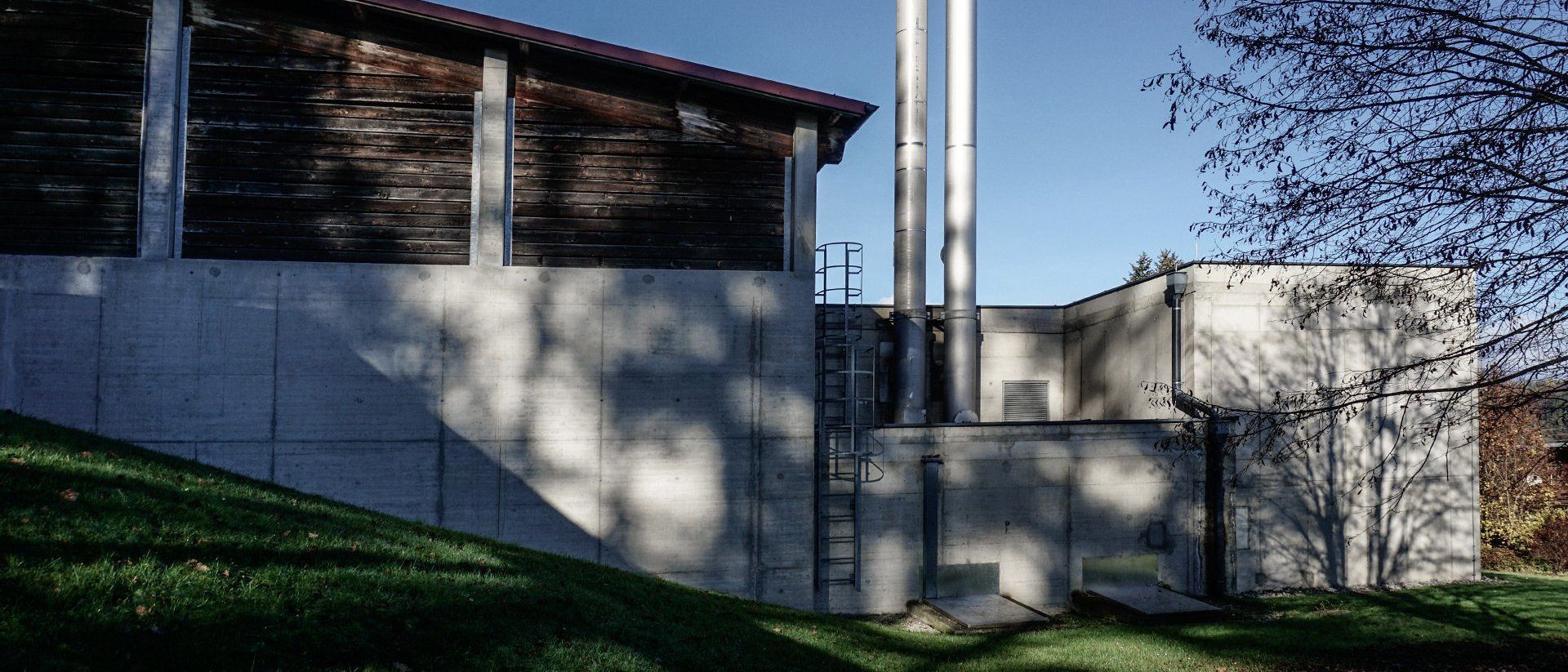 Ein modernes Gebäude mit einer braunen Holzfassade ist zu sehen, mit zwei hohen Schornsteinen und einer Betonwand. Die Szene ist im Freien mit einem klaren blauen Himmel im Hintergrund.