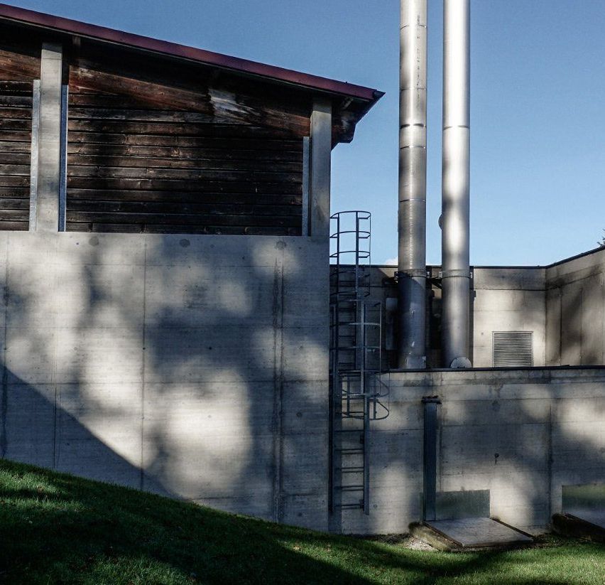 Ein modernes Gebäude mit einer braunen Holzfassade ist zu sehen, mit zwei hohen Schornsteinen und einer Betonwand. Die Szene ist im Freien mit einem klaren blauen Himmel im Hintergrund.