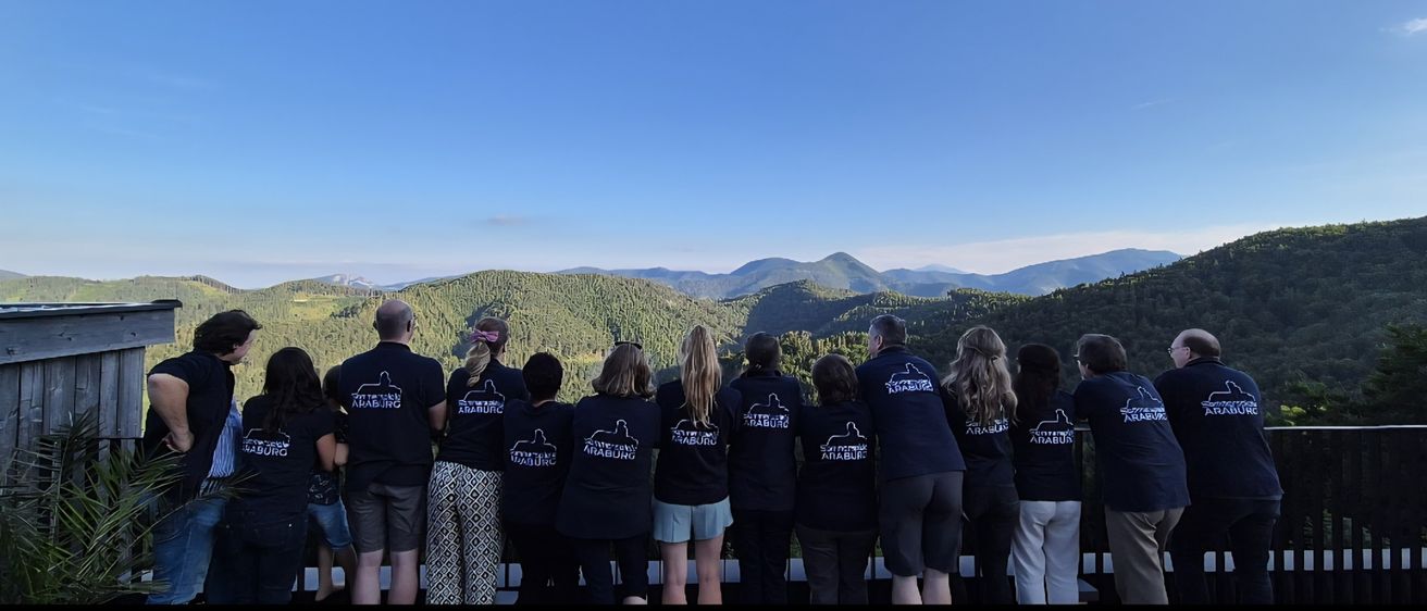 Eine Gruppe von Menschen in gleichen Shirts steht vor einem Zaun und blickt auf eine Bergkette mit klarem blauem Himmel.