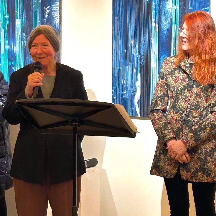 Zwei Frauen stehen vor einer weißen Wand. Eine hält ein Mikrofon, möglicherweise beim Sprechen, während die andere lächelt. Beide tragen Mäntel. Dahinter sind zwei abstrakte blaue Gemälde ausgestellt.