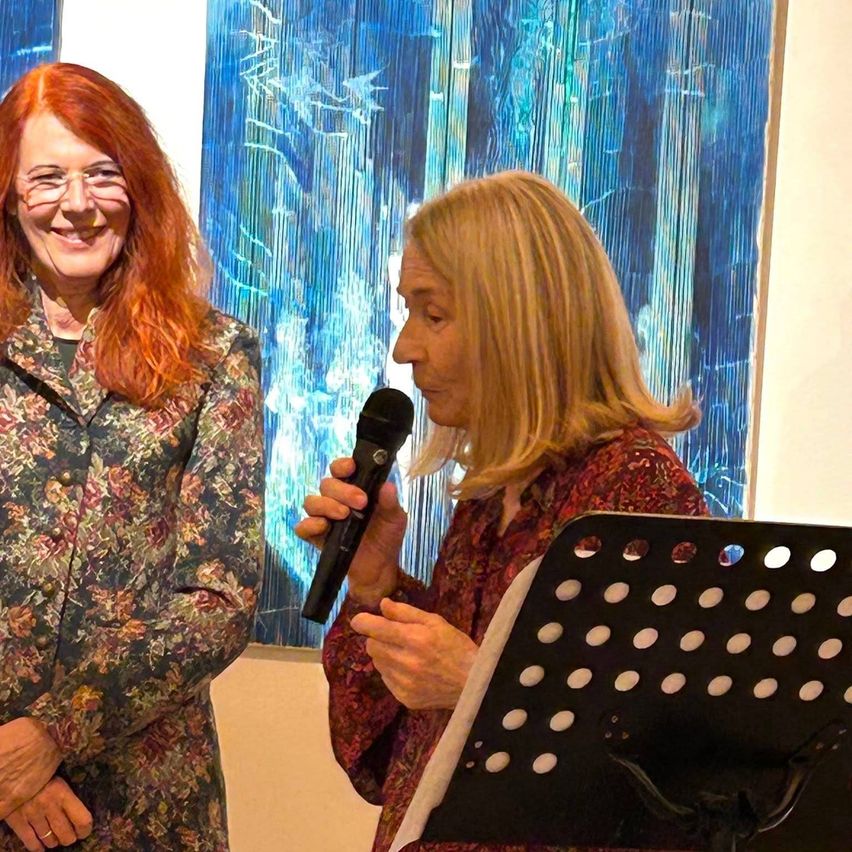 Zwei Frauen stehen vor einem blauen abstrakten Gemälde. Die Frau auf der rechten Seite hält ein Mikrofon, während die Frau auf der linken Seite lächelt.