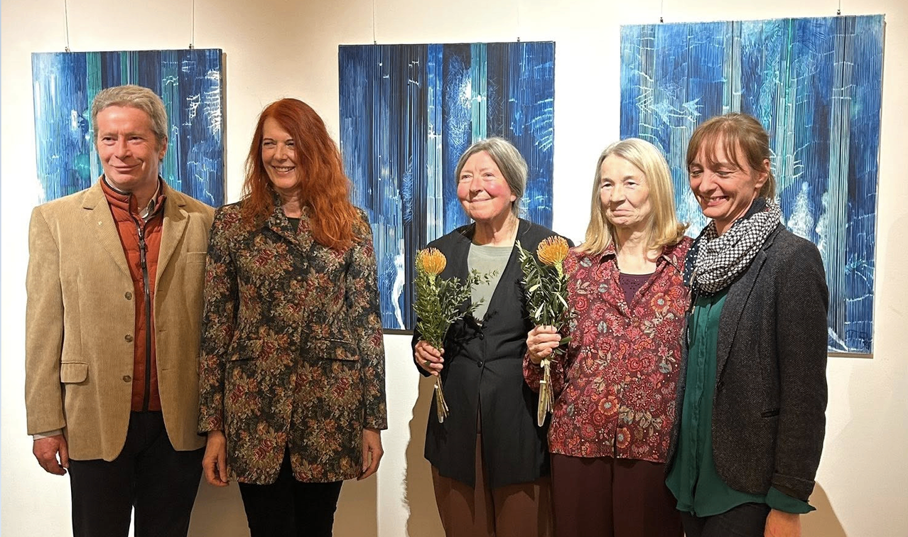 Fünf Frauen stehen eng beieinander vor blauen Gemälden und lächeln. Eine hält Blumen.