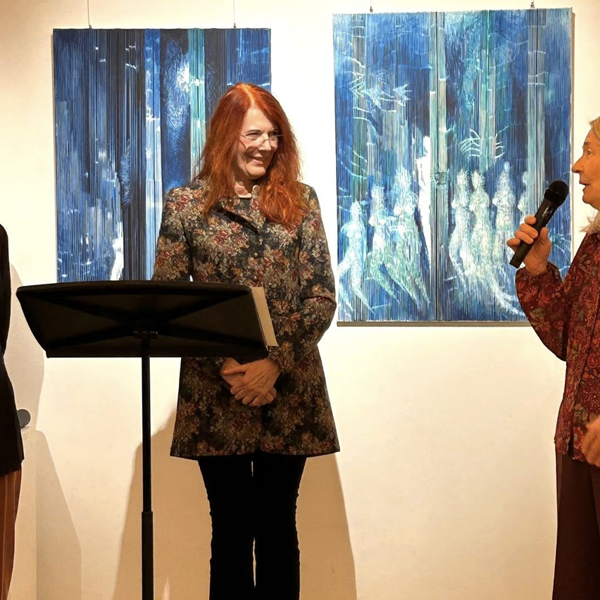 Zwei Frauen stehen vor einer Wand mit blauen Gemälden. Eine Frau hält ein Mikrofon und spricht, während die andere Frau zuhört und lächelt.