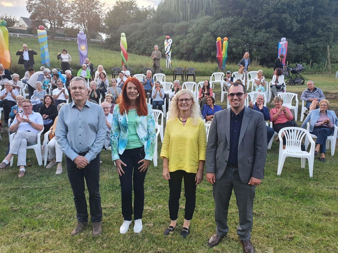 Vier Menschen stehen vor einer Menschenmenge in einem Außenbereich. Sie lächeln. Die Menge sitzt auf weißen Stühlen und beobachtet sie. Dahinter stehen farbenfrohe Skulpturen und Bäume.