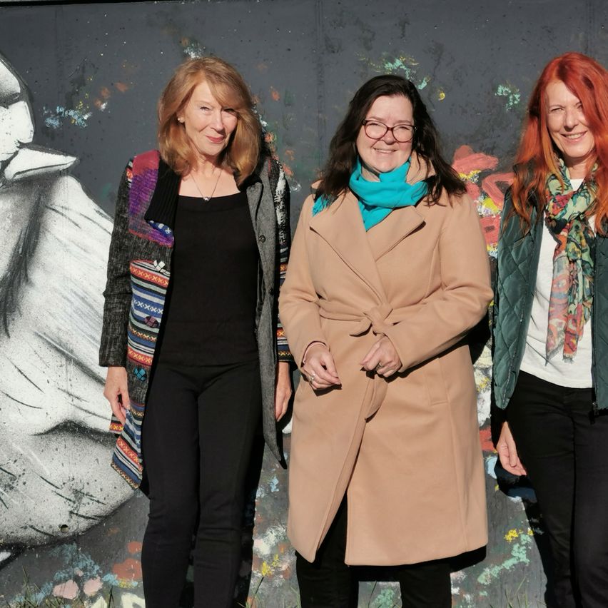 Drei Frauen stehen nebeneinander vor einer farbenfrohen Wandmalerei. Die Frau auf der linken Seite trägt ein schwarzes Oberteil und eine farbenfrohe Jacke. Die Frau in der Mitte trägt einen braunen Mantel und eine blaue Krawatte. Die Frau auf der rechten Seite trägt eine grüne Jacke und eine Blumenschal.