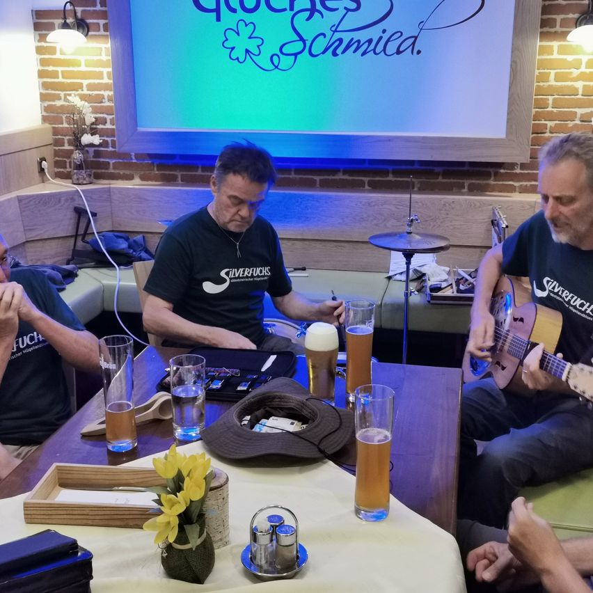 Drei Männer sitzen um einen Tisch mit Biergläsern herum. Einer spielt Gitarre, ein anderer Schlagzeug, und der dritte beobachtet. Eine Projektionsleinwand hinter ihnen zeigt Quockers Schmied.