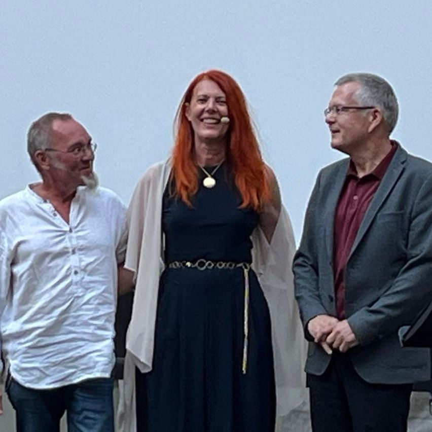 Drei Menschen stehen zusammen und lächeln. Die Frau trägt eine Halskette und ein Mikrofon am Ohr. Der Mann rechts trägt eine Brille und einen Anzug.