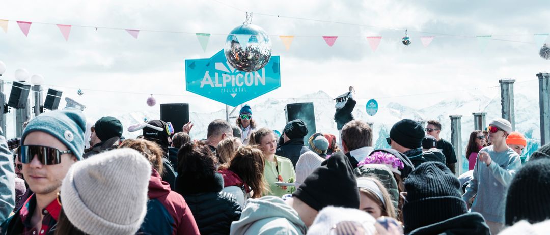 Eine Menschenmenge versammelt sich bei einer Outdoor-Veranstaltung, möglicherweise einem Musikfestival. Ein Schild mit der Aufschrift 'AlpiCon' hängt über ihnen, und eine Discokugel schwebt in der Luft. Eine Frau in einer grünen Jacke steht in der Mitte. Hinter ihr hält eine Person in einer weißen Maske ein Mikrofon. Schneebedeckte Berge bilden den Hintergrund.