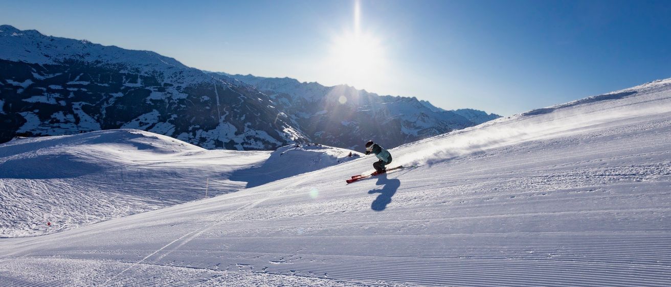 Ein Skifahrer fährt unter einem hellen Sonnenschein einen schneebedeckten Berghang hinunter, mit Bergen im Hintergrund.