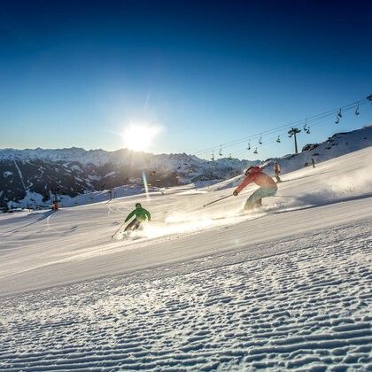 Zwei Skifahrer eilen unter einem strahlenden Sonnenschein einen schneebedeckten Berghang hinab. Sie sind von verschneiten Bergen und Skiliften umgeben.