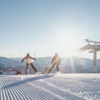 Zwei Skifahrer eilen einen verschneiten Berg hinunter, mit einer Skiliftstation im Hintergrund. Der Himmel ist hell und sonnig.