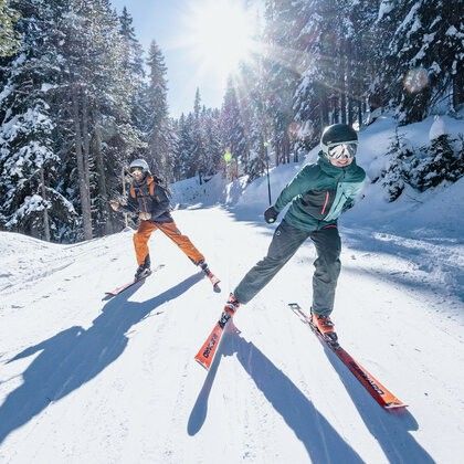 Zwei Skifahrer steigen an einem sonnigen Tag einen verschneiten Hang hinab und tragen Winterausrüstung.