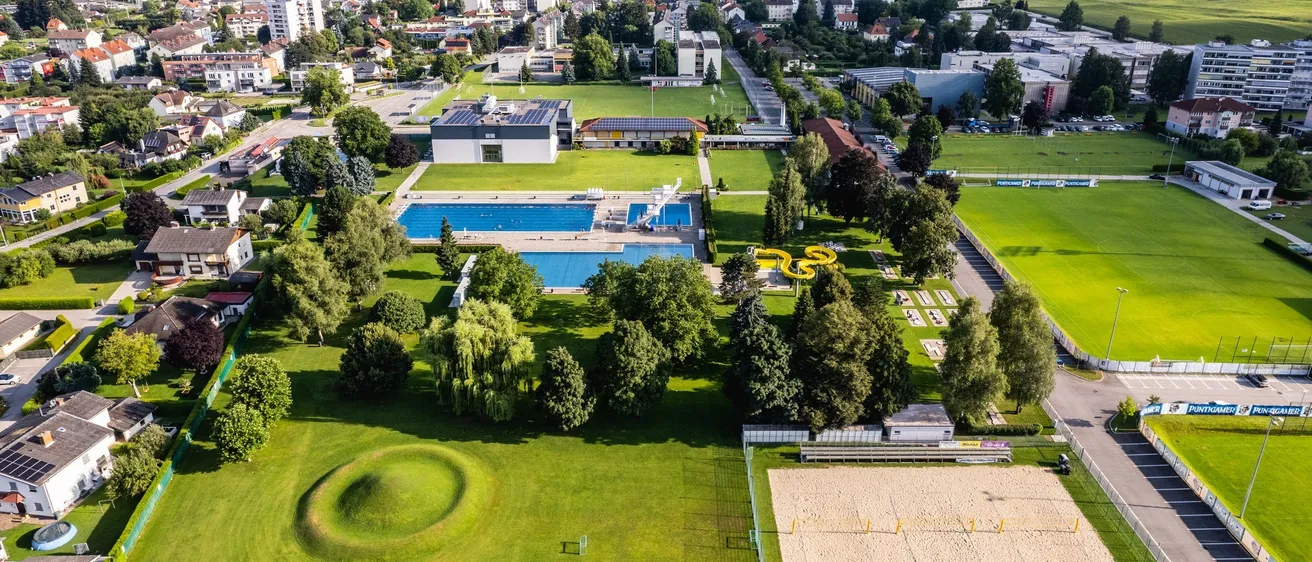 Luftaufnahme eines Parks mit Schwimmbad, Rutsche und Volleyballplatz, umgeben von Bäumen, Bergen und Gebäuden.