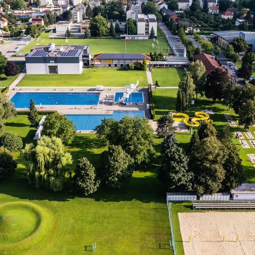 Luftaufnahme eines Parks mit Schwimmbad, Rutsche und Volleyballplatz, umgeben von Bäumen, Bergen und Gebäuden.