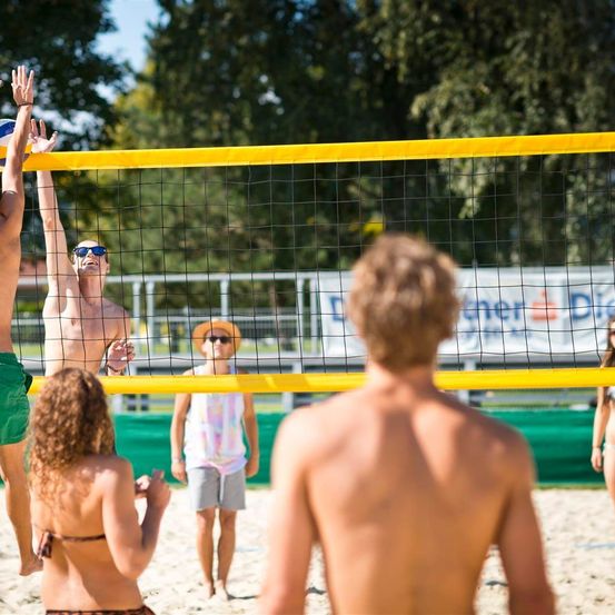 Eine Gruppe von Menschen spielt Beachvolleyball, ein Mann springt, um den Ball zu schlagen. Andere schauen im Hintergrund zu.