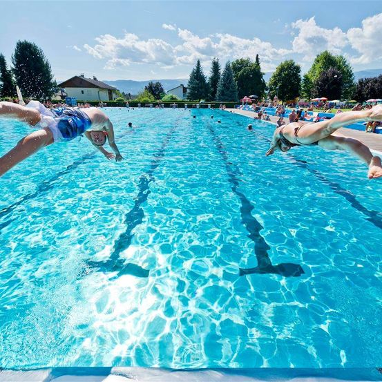Zwei Menschen springen an einem sonnigen Tag in einem Schwimmbad, viele Menschen sind in der Nähe.