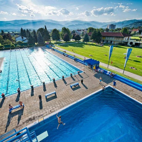 Ein Luftbild eines Freibades mit mehreren Schwimmenden und anderen Personen, die auf Bänken um den Pool herum sitzen. Der Pool ist von einem Rasenbereich mit Bäumen und Bergen in der Ferne umgeben.