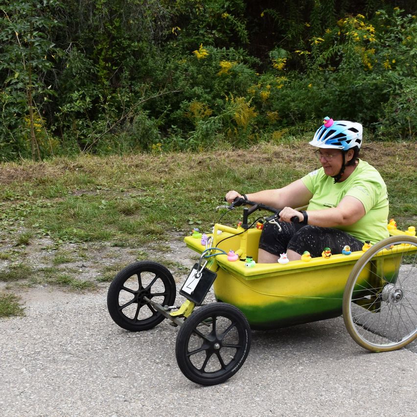 Eine Frau mit Helm fährt ein Dreirad mit einem gelben und grünen Eimer, gefüllt mit Gummienten. Sie ist auf einem Kiesweg mit viel Grün im Hintergrund.