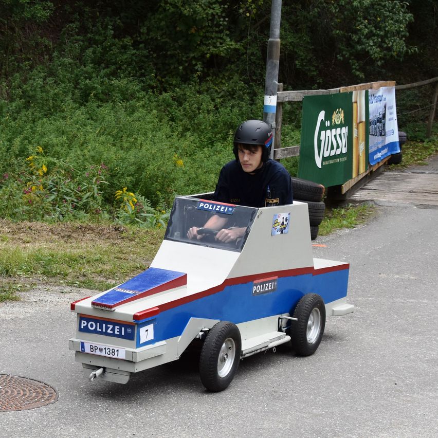 Eine Person mit Helm fährt ein kleines, blau-weißes Polizeiauto auf einer Straße. Das Auto hat das Wort 'POLIZEI' darauf.