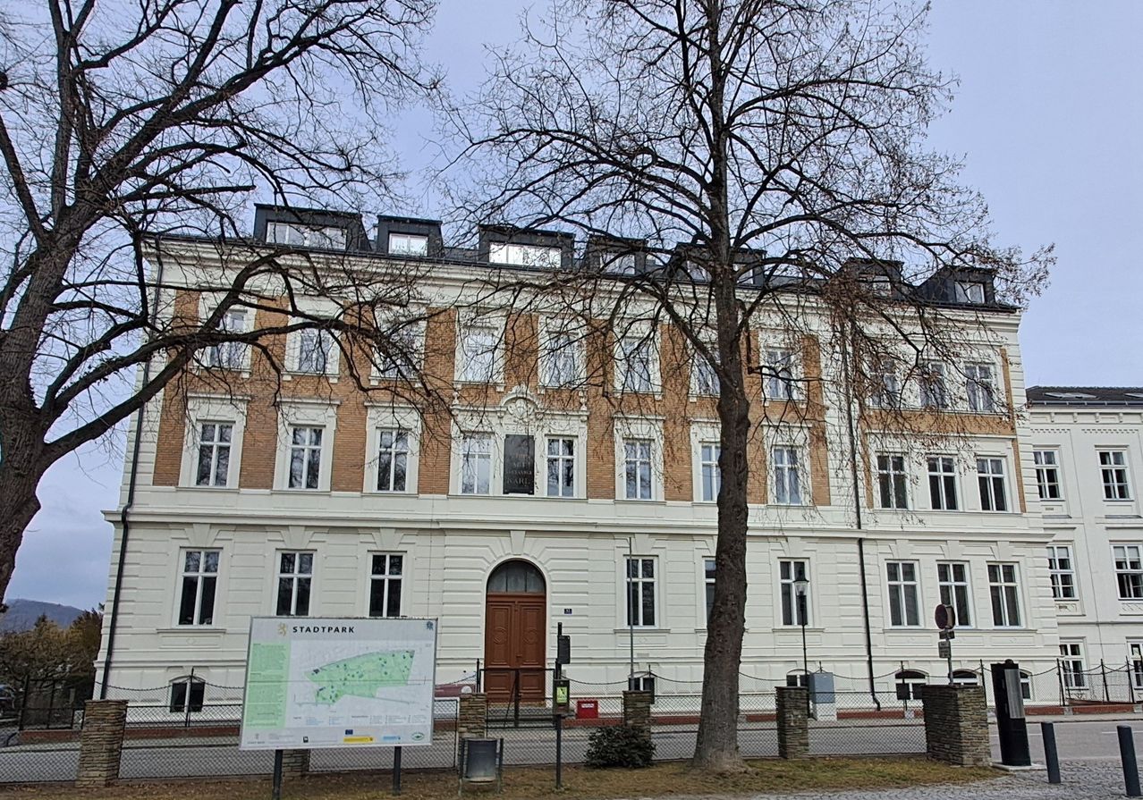 Ein großes mehrstöckiges Gebäude mit vielen Fenstern und einer braunen Tür ist von Bäumen und einem Zaun umgeben. Vor dem Gebäude ist eine Karte ausgestellt.