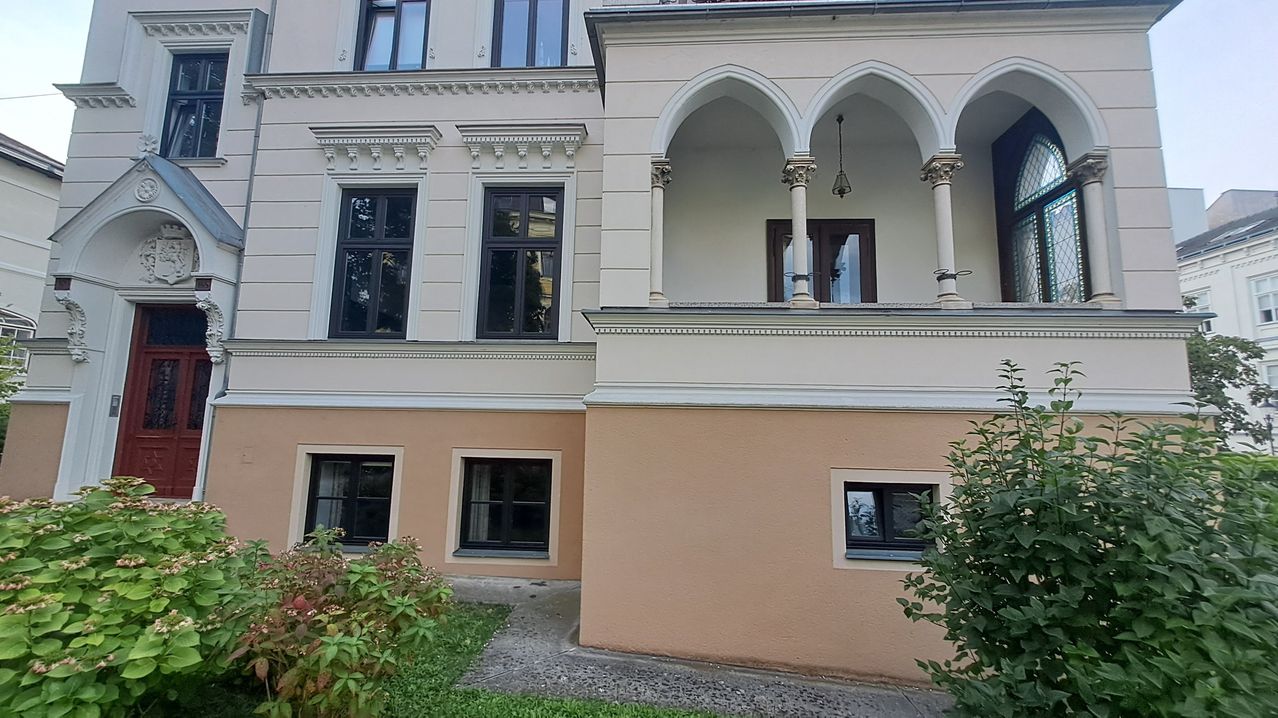 Das Bild zeigt die Fassade eines Gebäudes mit großen Bogenfenstern und einem Balkon. Die Wände sind in einem hellen Rosaton gestrichen, und es gibt dekorative Elemente an den Fenstern und Bögen.