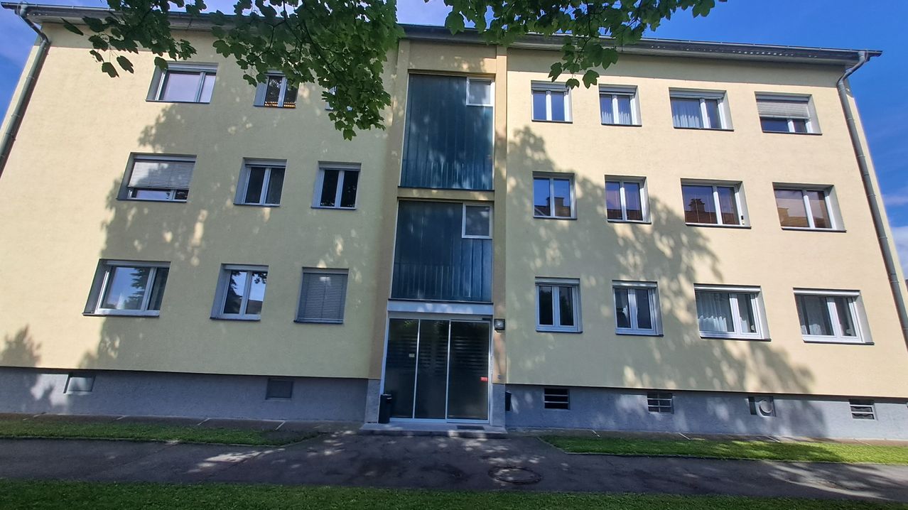 Ein gelbes Mehrfamilienhaus mit Glasfenstern und Türen, einem Mülleimer davor und einem Baum, der Schatten wirft.