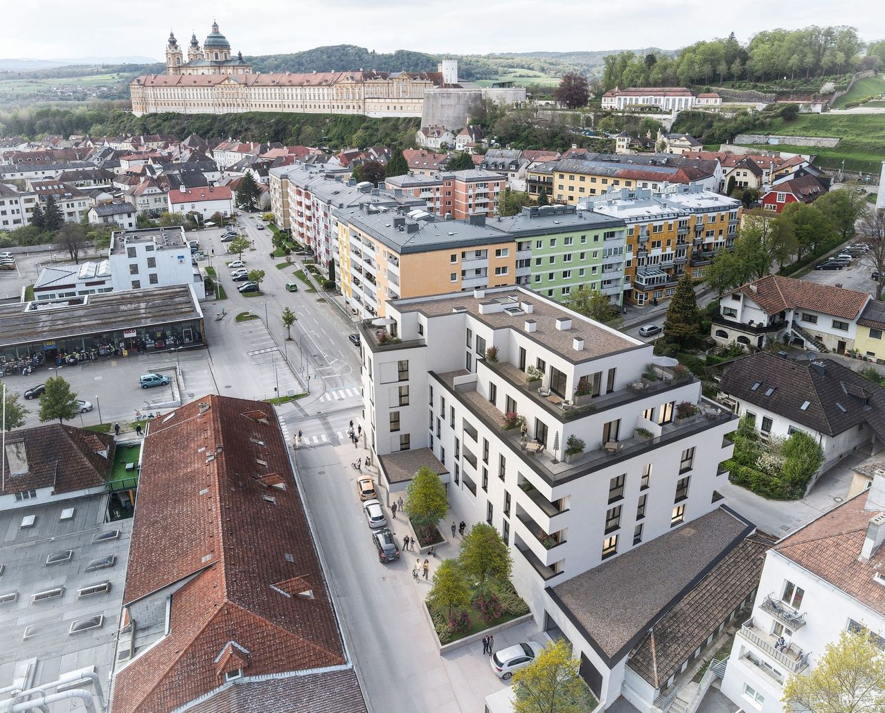 Luftaufnahme einer Stadt mit vielen Gebäuden, einige mit roten Dächern. Ein großes Schloss ist in der Ferne. Autos sind auf der Straße geparkt und Menschen gehen. Bäume und Pflanzen stehen vor den Gebäuden.