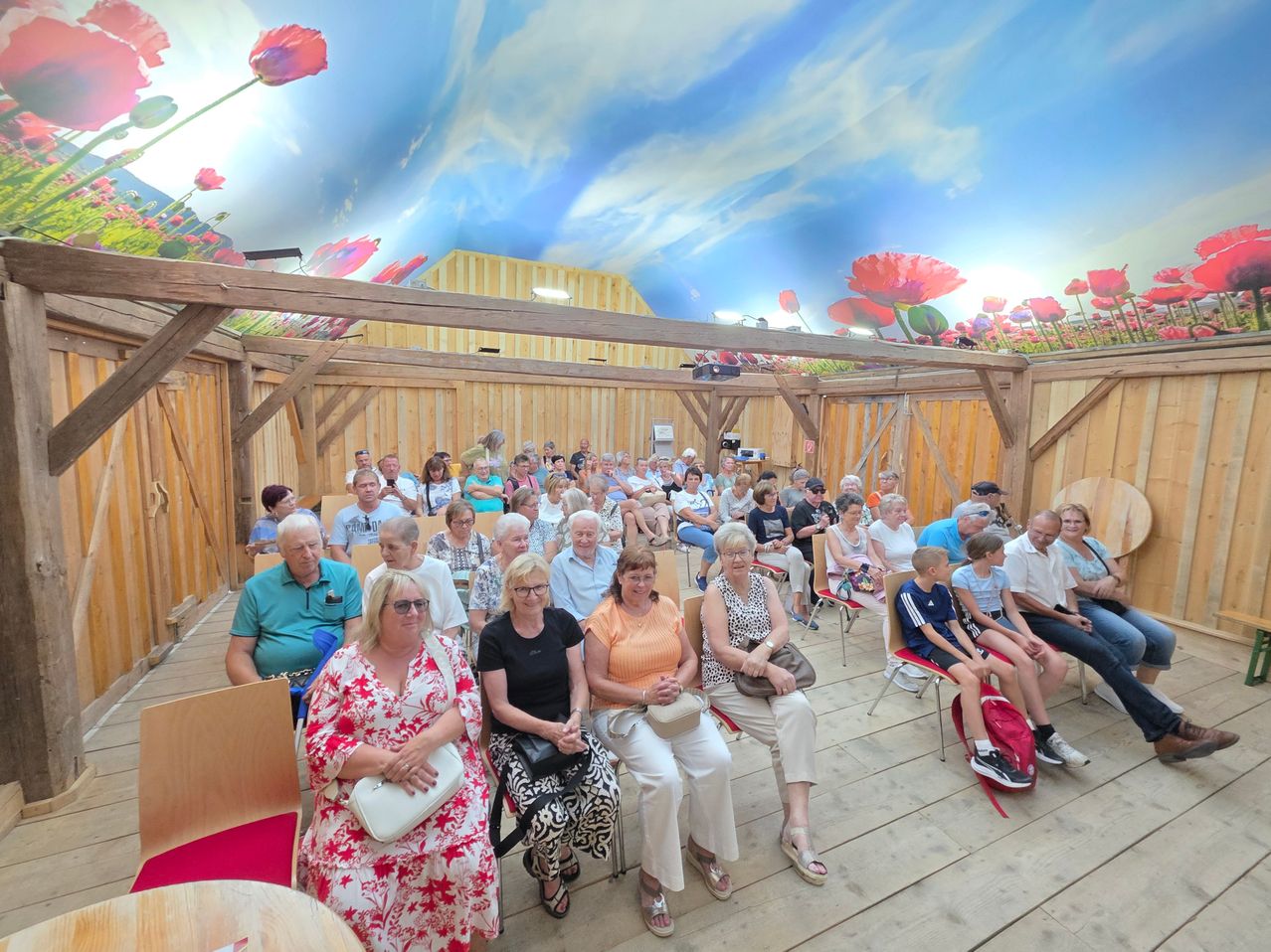 A group of people are sitting in a room with a wooden ceiling and walls painted with flowers. Some are wearing glasses and sneakers.
