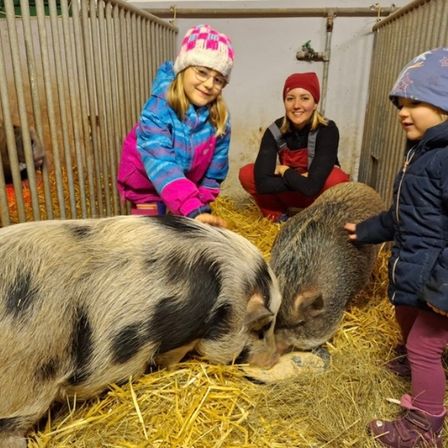 Drei Kinder, zwei Mädchen und eine Frau, sind in einer Scheune und füttern zwei Schweine. Die Frau sitzt auf dem Boden, und die Mädchen stehen neben den Schweinen, von denen eines etwas vom Boden frisst.