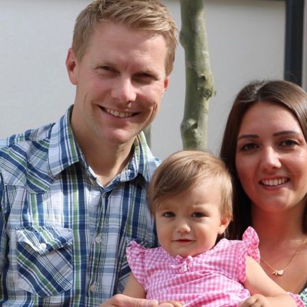 Eine dreiköpfige Familie lächelt für ein Foto. Der Mann trägt ein kariertes Hemd, die Frau ein rosa Kleid, und das Baby sitzt zwischen ihnen und lächelt ebenfalls.