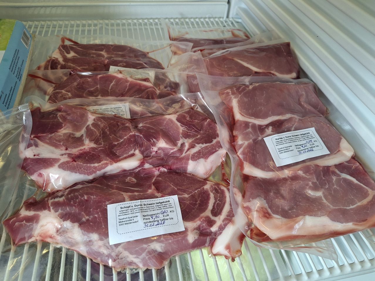 Mehrere Stücke rohes Schweinefleisch sind in einem Kühlschrank ausgestellt, jedes in seiner eigenen Plastiktüte mit weißen Etiketten, die Informationen enthalten. Das Schweinefleisch hat eine rosa-rote Farbe und ist frisch.