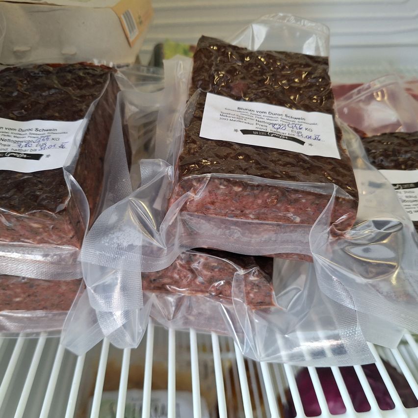 Mehrere Packungen mit Hackfleisch sind in Plastik verpackt und in einem Kühlschrank angeordnet. Jede Packung hat ein weißes Etikett mit schwarzem Text. Ein Etikett lautet 'Blunzen vom Duroc Schwein'. Das Fleisch ist braun und leicht glänzend.