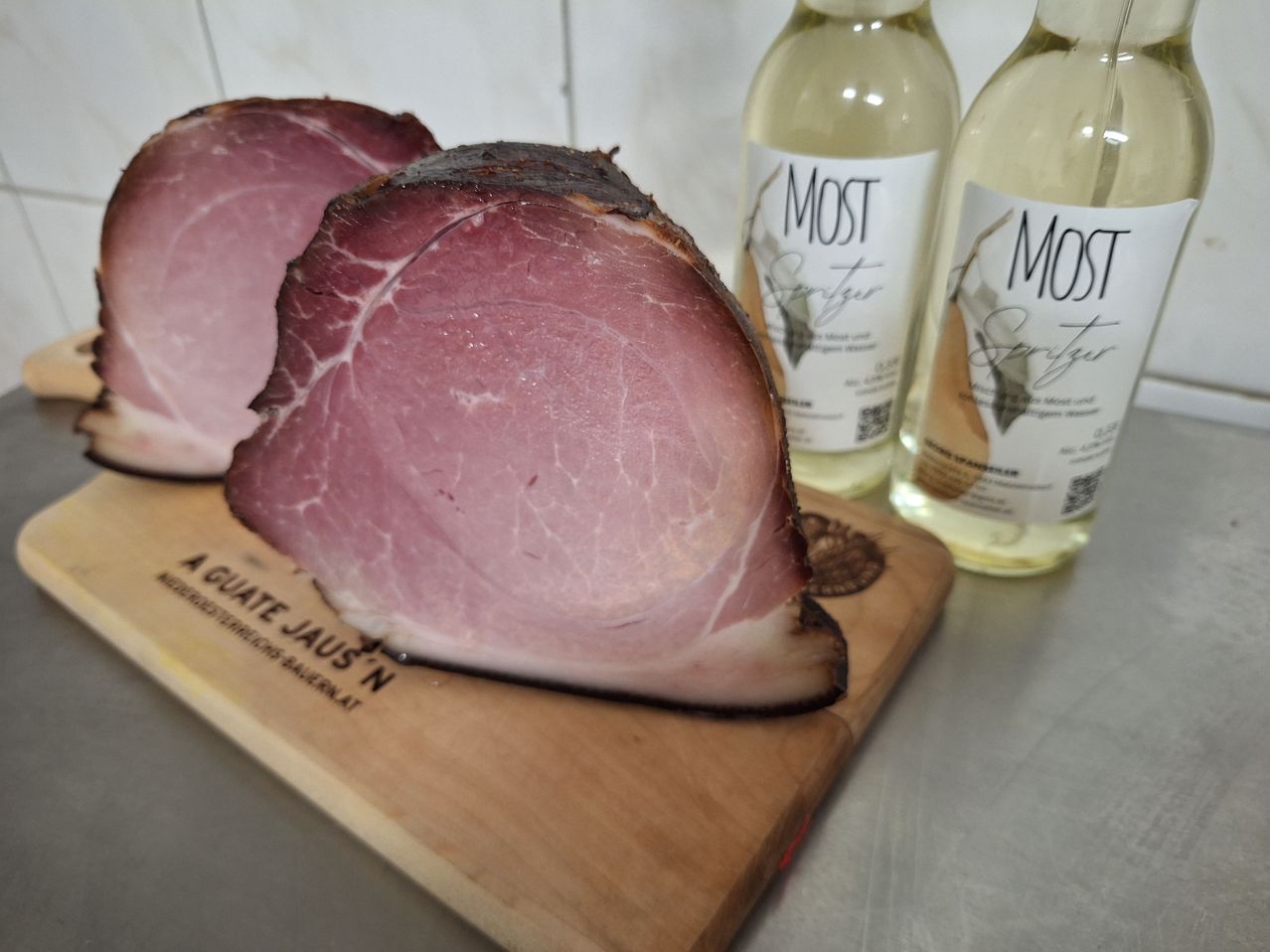 Ein geschnittener Schinken liegt auf einem Holzschneidebrett neben zwei Flaschen Weißwein mit der Aufschrift 'MOST'.