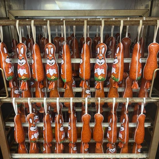 A row of orange Easter sausages with bunny faces hang on a metal rack, each with a red bow tie and green grass.