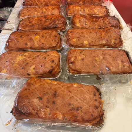 Mehrere Meatloaf-Laibe sind in einer Reihe angeordnet, jeweils in Plastik eingewickelt und in Aluminiumfolien-Schalen auf einer weißen Oberfläche platziert.