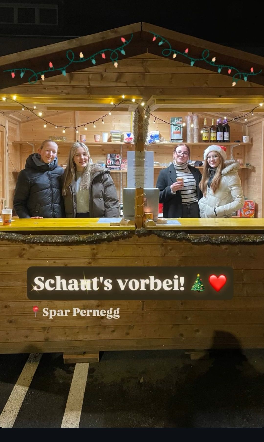 Vier Frauen stehen hinter einer Theke in einem chaletähnlichen Holzhäuschen, geschmückt mit Weihnachtsdekoration. Eine hält ein Getränk, eine andere trägt einen Weihnachtsmannhut. Die Theke trägt den Schriftzug 'Schaut's vorbei!' mit einem Weihnachtsbaum und Herz-Emoji.