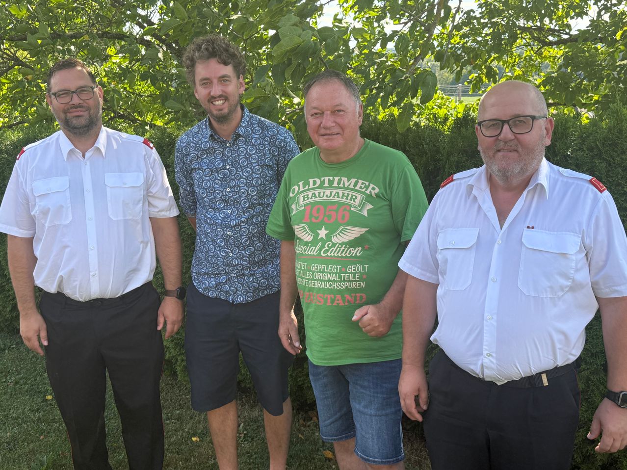 Vier Männer stehen in einem Garten und lächeln. Der Mann auf der linken Seite trägt ein weißes Hemd und schwarze Hose. Der zweite Mann von links trägt ein blaues Hemd mit Muster und dunkle Shorts. Der dritte Mann trägt ein grünes Hemd mit dem Text 'Oldtimer Baujahr 1956' und blaue Jeans. Der Mann auf der rechten Seite trägt ein weißes Hemd und schwarze Hose.