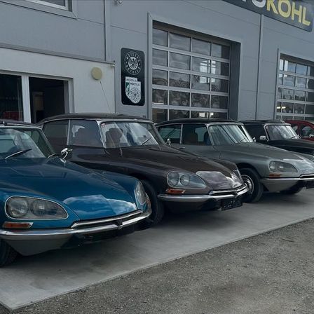 Fünf Oldtimer, darunter ein blauer, schwarzer, grauer und roter Citroen DS, sind vor einem Gebäude mit dem Wort Kohl oben geparkt.