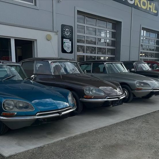 Fünf Oldtimer, darunter ein blauer, schwarzer, grauer und roter Citroen DS, sind vor einem Gebäude mit dem Wort Kohl oben geparkt.