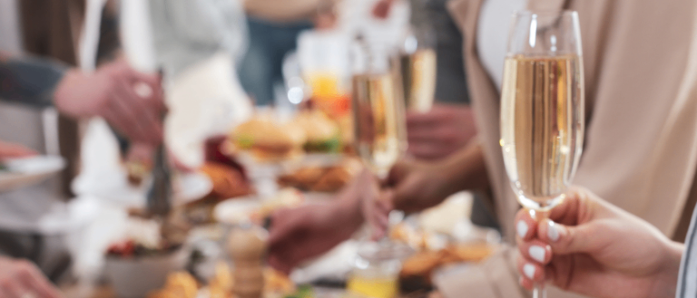 Eine Gruppe von Menschen an einem Buffet-Tisch mit verschiedenen Speisen, einer hält einen Teller mit Brot und Tomaten, ein anderer eine Champagnerflöte.